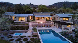 A residence emits warm light through its windows and doors. It is surrounded by views of desert foliage and a pool.