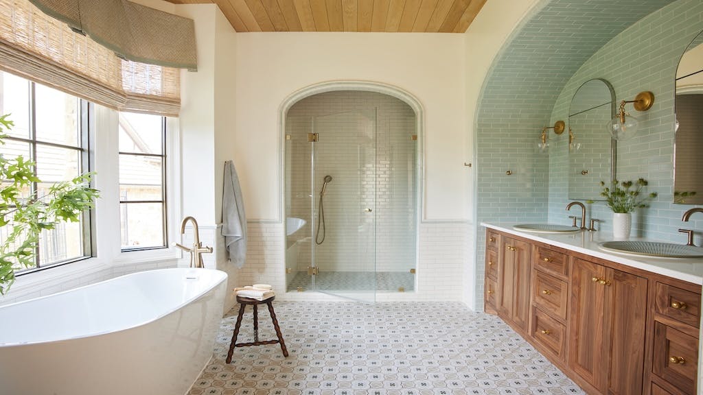 A large bathroom with freestanding tub underneath windows to the left and double wooden vanity to the right. In the back of the room is a shower with an arched entryway.