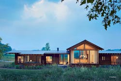 A single-story timber frame home glows through large windows as it sits on a rural hillside. A single-story timber frame home glows through large windows as it sits on a rural hillside.